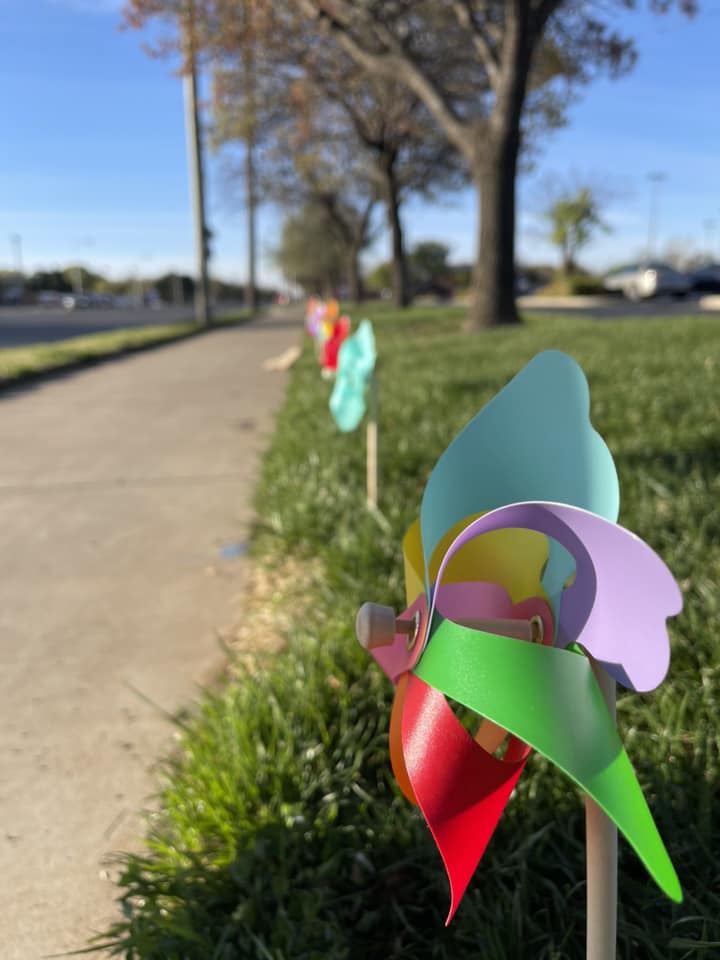 Colorful pinwheels lined up along a sidewalk in green grass on a sunny day