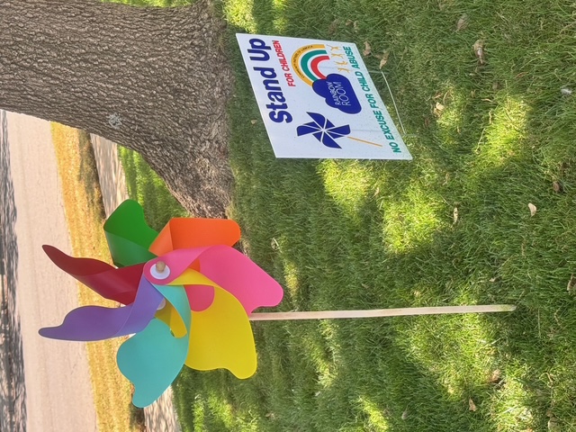A large rainbow pinwheel next to a Stand Up For Children yard sign under a tree